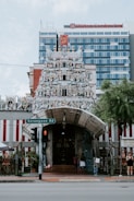 The image depicts a richly adorned Hindu temple facade with numerous intricately detailed sculptures of deities and mythological figures. The temple has a grand entrance with a tall gopuram, and the vibrant colors and decorations stand out. Behind the temple, there is a modern multistory building, and in the foreground, there is a street sign for Serangoon Road and a traffic light. A few people are near the entrance.