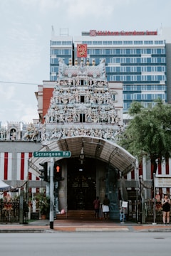The image depicts a richly adorned Hindu temple facade with numerous intricately detailed sculptures of deities and mythological figures. The temple has a grand entrance with a tall gopuram, and the vibrant colors and decorations stand out. Behind the temple, there is a modern multistory building, and in the foreground, there is a street sign for Serangoon Road and a traffic light. A few people are near the entrance.