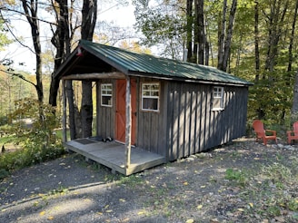 A rustic wooden cabin with a green metal roof is situated in a wooded area surrounded by tall trees. The cabin has a small porch with a few steps leading up to a door painted in a warm brown-orange color. Two small windows are visible on the front of the cabin. Nearby, two red Adirondack chairs are placed on the gravel ground, suggesting a seating area for relaxation.