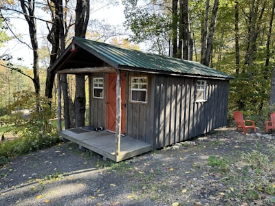 A rustic wooden cabin with a green metal roof is situated in a wooded area surrounded by tall trees. The cabin has a small porch with a few steps leading up to a door painted in a warm brown-orange color. Two small windows are visible on the front of the cabin. Nearby, two red Adirondack chairs are placed on the gravel ground, suggesting a seating area for relaxation.