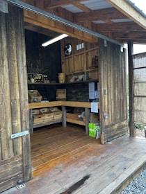 A rustic wooden shed with open sliding doors reveals shelves filled with baskets and wooden crates containing various items. A small display offers goods like honey, and informational flyers are pinned to the walls. A light hangs from the ceiling, illuminating the interior.