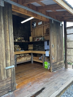 A rustic wooden shed with open sliding doors reveals shelves filled with baskets and wooden crates containing various items. A small display offers goods like honey, and informational flyers are pinned to the walls. A light hangs from the ceiling, illuminating the interior.