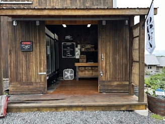 A rustic wooden structure serves as a small shop or market stall. The interior is visible through large open doors, revealing shelves with boxes and a fan. A blackboard sign displays a menu or list of products. A sign reading 'OPEN' is mounted on the left, and a flag with the same word hangs on the right. The exterior area is surrounded by gravel and a barrel planter with flowers.