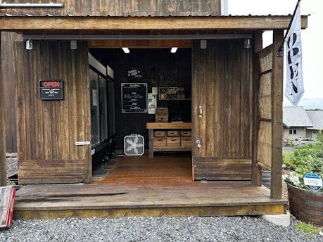 A rustic wooden structure serves as a small shop or market stall. The interior is visible through large open doors, revealing shelves with boxes and a fan. A blackboard sign displays a menu or list of products. A sign reading 'OPEN' is mounted on the left, and a flag with the same word hangs on the right. The exterior area is surrounded by gravel and a barrel planter with flowers.