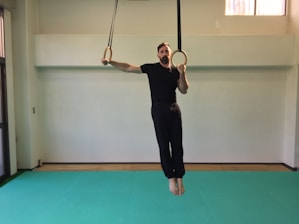 A man dressed in black is holding gymnastics rings suspended from the ceiling in a brightly lit room. The floor is covered with a light green mat, and there are windows providing natural light. The walls are plain and light-colored.
