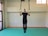 A person is performing an exercise on gymnastic rings, holding themselves steady while wearing black athletic clothing. The setting appears to be an indoor gym with a padded floor, and natural light is coming through a window on the side.