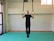 A person is performing an exercise on gymnastic rings, holding themselves steady while wearing black athletic clothing. The setting appears to be an indoor gym with a padded floor, and natural light is coming through a window on the side.