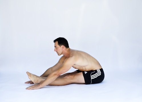 A man with short dark hair is sitting on the floor in a forward bend position, reaching for his toes. He is shirtless and wearing black shorts with white stripes. The background is plain and light-colored, emphasizing the focus on the individual's stretching pose.