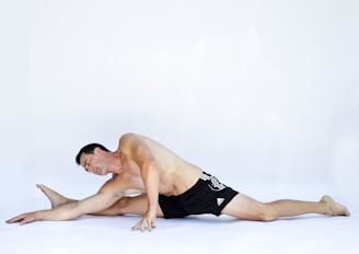 A man is performing a seated forward stretch, with one leg extended to the side and the other bent. He is shirtless and wearing black athletic shorts, positioned on a white background.
