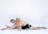 A man is performing a seated forward stretch, with one leg extended to the side and the other bent. He is shirtless and wearing black athletic shorts, positioned on a white background.