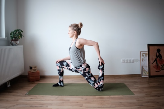A person is performing a yoga or stretching pose on a green yoga mat in a minimalist room with wooden flooring. They are balancing on one knee while holding the other foot with one hand. The person is wearing patterned leggings and a sleeveless gray top. A small plant on a radiator, stacked blocks, and framed artwork are visible in the background.