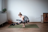 A person is practicing yoga on a green mat in a minimalistic room. The individual is balancing on their hands with legs crossed off the ground, showcasing strength and flexibility. The room features hardwood floors, a potted plant on a radiator, yoga blocks, and framed artwork leaning against the wall.