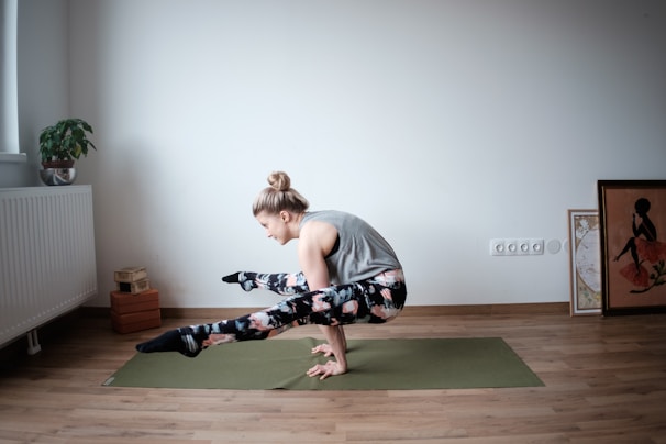 A person is practicing yoga on a green mat in a minimalistic room. The individual is balancing on their hands with legs crossed off the ground, showcasing strength and flexibility. The room features hardwood floors, a potted plant on a radiator, yoga blocks, and framed artwork leaning against the wall.