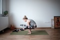 A person is practicing yoga on a green mat in a minimalistic room. The individual is balancing on their hands with legs crossed off the ground, showcasing strength and flexibility. The room features hardwood floors, a potted plant on a radiator, yoga blocks, and framed artwork leaning against the wall.
