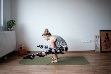 A person is practicing yoga on a green mat in a minimalistic room. The individual is balancing on their hands with legs crossed off the ground, showcasing strength and flexibility. The room features hardwood floors, a potted plant on a radiator, yoga blocks, and framed artwork leaning against the wall.