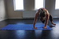 A person is performing a yoga or exercise pose on a blue mat in a well-lit room. The individual is in a plank position, balancing on their hands and toes, with one knee bent and pulled towards the chest. The room has neutral-colored walls and large windows, allowing natural light to illuminate the space.