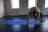 A person is performing a yoga or exercise pose on a blue mat in a well-lit room. The individual is in a plank position, balancing on their hands and toes, with one knee bent and pulled towards the chest. The room has neutral-colored walls and large windows, allowing natural light to illuminate the space.
