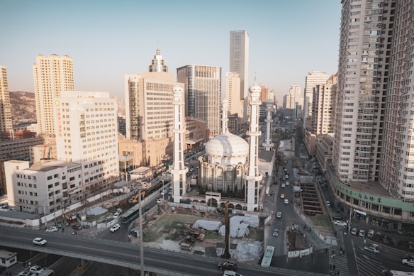 A bustling urban landscape with a large, white domed mosque situated in the center, surrounded by tall modern skyscrapers. The scene captures a busy city street with various vehicles and construction activity around the mosque.