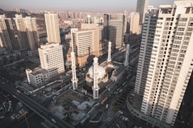 An aerial view of a densely built urban area featuring a mix of high-rise buildings and a mosque with four prominent minarets. The mosque's dome and minarets stand out among the modern skyscrapers, indicating a blend of traditional and contemporary architecture. The streets and city infrastructure are visible, including roads with cars and buses, reflecting a bustling city life.