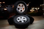 Three stacked car tires are placed on a concrete surface. Each tire has a shiny silver rim. The scene is set outdoors at night, with faint illumination from decorative lights in the background. A black vehicle is partially visible to the left.