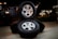 Three stacked car tires are placed on a concrete surface. Each tire has a shiny silver rim. The scene is set outdoors at night, with faint illumination from decorative lights in the background. A black vehicle is partially visible to the left.
