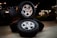 Three stacked car tires are placed on a concrete surface. Each tire has a shiny silver rim. The scene is set outdoors at night, with faint illumination from decorative lights in the background. A black vehicle is partially visible to the left.