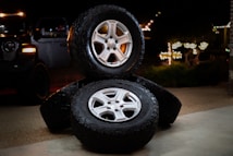 Three stacked car tires are placed on a concrete surface. Each tire has a shiny silver rim. The scene is set outdoors at night, with faint illumination from decorative lights in the background. A black vehicle is partially visible to the left.