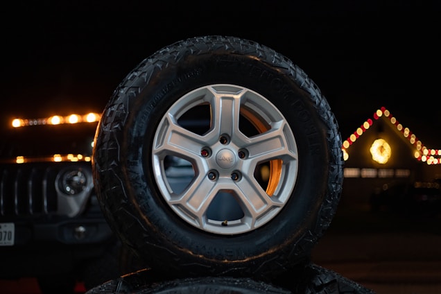 A Jeep tire is prominently displayed in the foreground with intricate tread patterns. In the background, a vehicle with illuminated headlights is partially visible, and festive lights adorn a roofline, creating a warm and cheerful atmosphere.