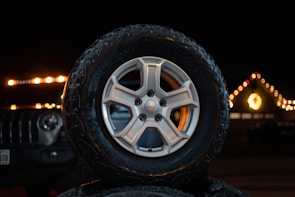 A Jeep tire is prominently displayed in the foreground with intricate tread patterns. In the background, a vehicle with illuminated headlights is partially visible, and festive lights adorn a roofline, creating a warm and cheerful atmosphere.