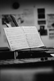 A black and white photograph of an open music book on a stand in a dimly lit room. The background is blurred but shows some wall posters and frames. The focus is on the sheet music, highlighting musical notes and classical arrangements.