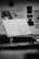 A black and white photograph of an open music book on a stand in a dimly lit room. The background is blurred but shows some wall posters and frames. The focus is on the sheet music, highlighting musical notes and classical arrangements.