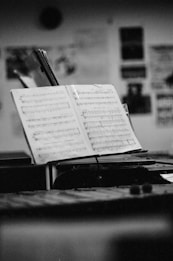 A black and white photograph of an open music book on a stand in a dimly lit room. The background is blurred but shows some wall posters and frames. The focus is on the sheet music, highlighting musical notes and classical arrangements.