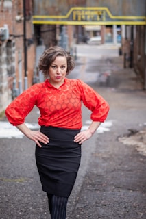 A person stands confidently in an alleyway, wearing a bold red top and a black skirt. The background features urban elements such as brick walls and a metallic sign with yellow accents. The pose conveys a sense of self-assurance.
