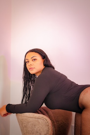 A woman with long, dark hair is leaning on a cushioned chair. She is wearing a black outfit and fishnet stockings, set against a plain, light-colored background.