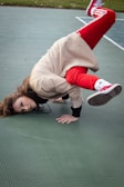 A person wearing a beige coat, red pants, red sneakers, and white socks is performing an acrobatic or breakdance move on a greenish floor. The individual is balancing on one hand with their head close to the ground while the legs are elevated, demonstrating agility and skill.