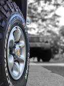 A close-up view of a vehicle tire with the brand name Cooper and model Discoverer AT3 XLT. The tire's treads and grooves are clearly visible, with a shiny metallic hubcap at the center. The background appears to be blurred, featuring a larger vehicle possibly parked or in motion.