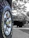 A close-up view of a vehicle tire with the brand name Cooper and model Discoverer AT3 XLT. The tire's treads and grooves are clearly visible, with a shiny metallic hubcap at the center. The background appears to be blurred, featuring a larger vehicle possibly parked or in motion.