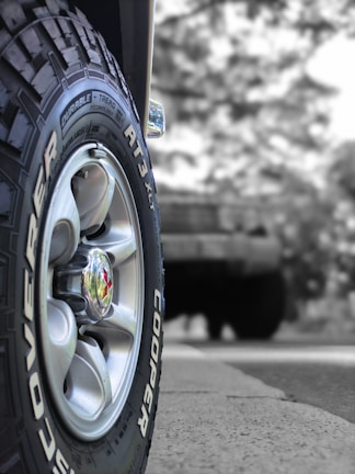 A close-up view of a vehicle tire with the brand name Cooper and model Discoverer AT3 XLT. The tire's treads and grooves are clearly visible, with a shiny metallic hubcap at the center. The background appears to be blurred, featuring a larger vehicle possibly parked or in motion.