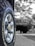 A close-up view of a vehicle tire with the brand name Cooper and model Discoverer AT3 XLT. The tire's treads and grooves are clearly visible, with a shiny metallic hubcap at the center. The background appears to be blurred, featuring a larger vehicle possibly parked or in motion.
