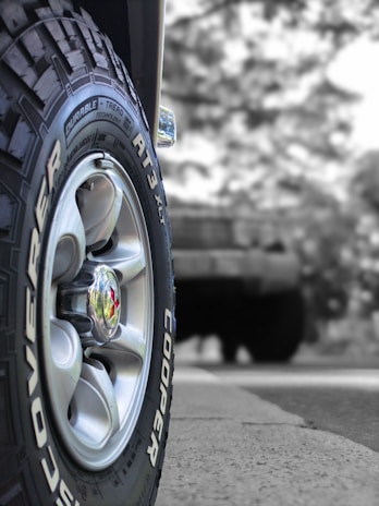 A close-up view of a vehicle tire with the brand name Cooper and model Discoverer AT3 XLT. The tire's treads and grooves are clearly visible, with a shiny metallic hubcap at the center. The background appears to be blurred, featuring a larger vehicle possibly parked or in motion.