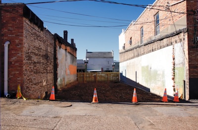An empty urban plot located between two old brick buildings. The area is surrounded by orange traffic cones and caution tape, indicating possible construction or maintenance activity. The ground appears to be freshly cleared earth. Overhead, several power lines stretch across the clear blue sky. A small wooden fence is at the far end of the plot, separating it from another building visible in the background.