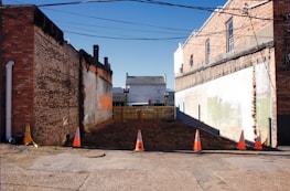 An empty urban plot located between two old brick buildings. The area is surrounded by orange traffic cones and caution tape, indicating possible construction or maintenance activity. The ground appears to be freshly cleared earth. Overhead, several power lines stretch across the clear blue sky. A small wooden fence is at the far end of the plot, separating it from another building visible in the background.