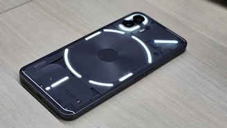 A smartphone displaying a transparent back panel with visible internal components, highlighted by a series of bright white LED strips. The device rests on a wooden surface, and the dual camera module is prominently visible.