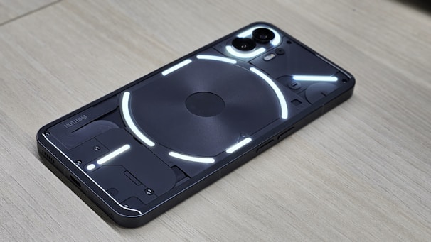 A smartphone displaying a transparent back panel with visible internal components, highlighted by a series of bright white LED strips. The device rests on a wooden surface, and the dual camera module is prominently visible.