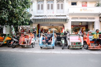 A row of colorful rickshaws is parked on a street in front of a building with multiple shops. Several men are seated in the rickshaws, looking relaxed and casual. The trees on the left add a touch of greenery, while the shops behind display various products, creating a bustling street atmosphere.