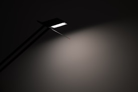 A minimalist lamp casts a soft white light onto a plain wall, highlighting a sleek, linear design with a sharp, angular edge. The lamp is obscured partially in shadow, emphasizing the contrast between light and dark.