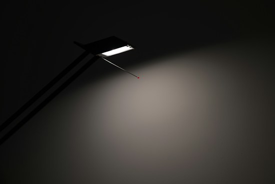 A minimalist lamp casts a soft white light onto a plain wall, highlighting a sleek, linear design with a sharp, angular edge. The lamp is obscured partially in shadow, emphasizing the contrast between light and dark.