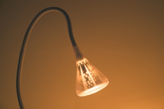 A glowing lamp with a curved neck emits a warm light. The lamp's translucent shade is illuminated from within, creating a cozy ambiance against a minimalistic background.