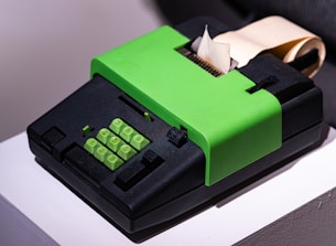 A retro-style adding machine with a bright green cover and keypad. It features a numeric keypad and some function keys. A roll of paper is partially extended behind it.