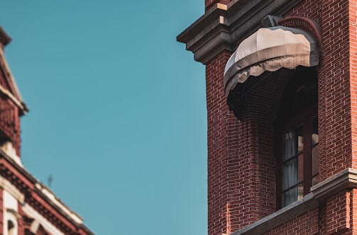 A close-up view of a red brick building with an ornate architectural style. A window with an arched design is partially covered by a beige fabric awning, casting shadows on the wall. The building features detailed brickwork with a classic cornice. The sky in the background is clear and blue, adding contrast to the warm tones of the building.